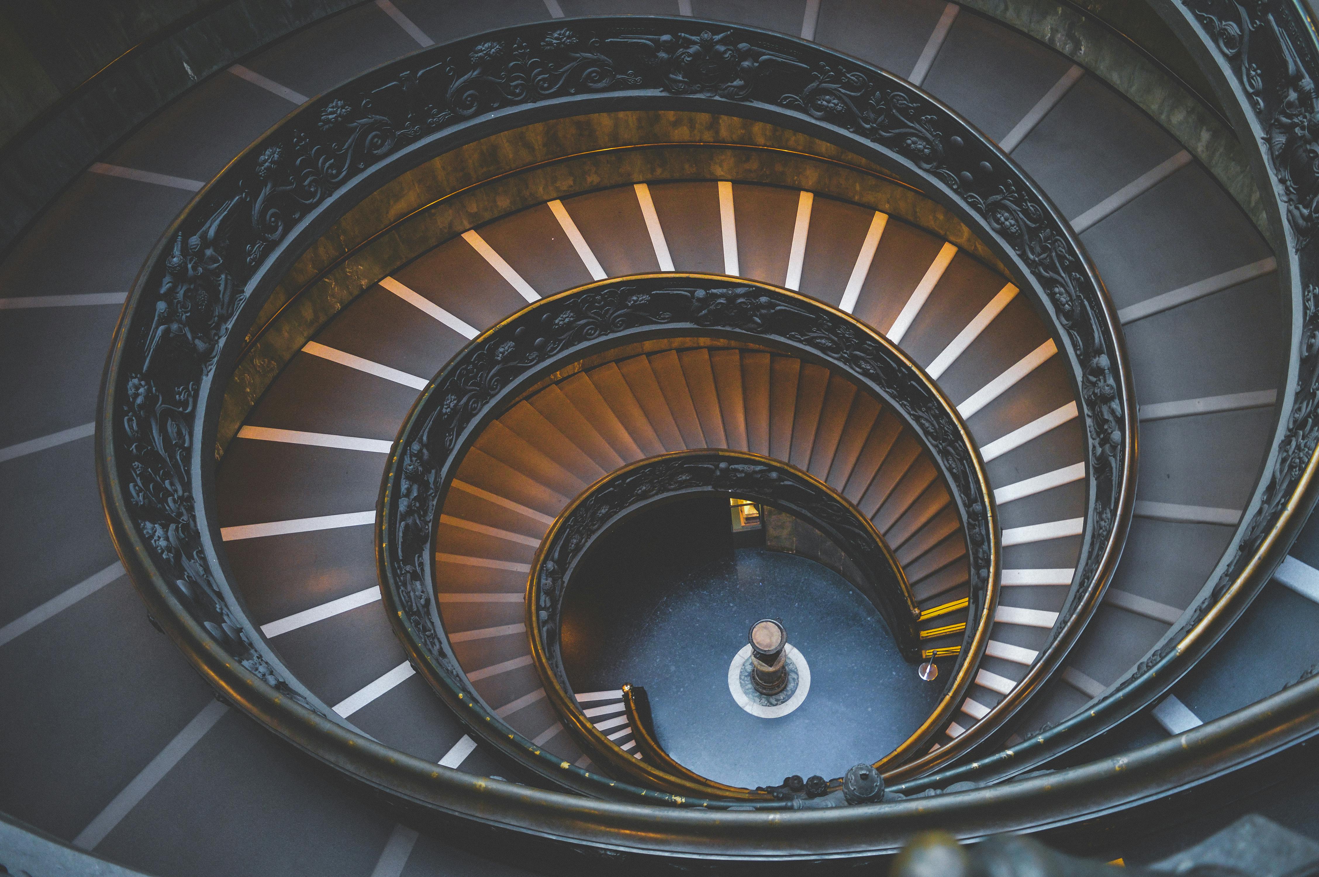 Ornate spiral staircase seen from above, dark ironwork and warm steps, suggesting focus, calm, and a reflective journey.